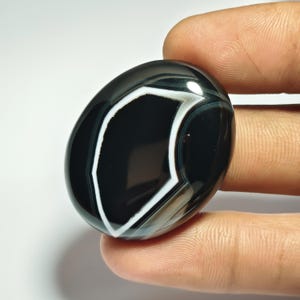 May include: A polished, oval-shaped gemstone with a striking black and white banded pattern. The stone features a prominent white, angular design against a glossy black background. The gemstone is held between fingers, showcasing its smooth, reflective surface.
