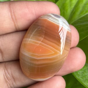 May include: Oval-shaped polished gemstone with bands of orange, white, and brown. The stone is held in a hand, showcasing its smooth, glossy surface. The gemstone's natural patterns create a unique, eye-catching design. This item is perfect for jewelry making or collecting.