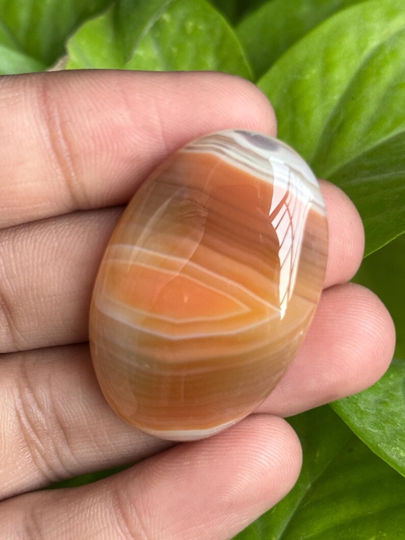 May include: Oval-shaped polished gemstone with bands of orange, white, and brown. The stone is held in a hand, showcasing its smooth, glossy surface. The gemstone's natural patterns create a unique, eye-catching design. This item is perfect for jewelry making or collecting.
