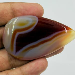 May include: A polished, teardrop-shaped cabochon gemstone featuring smooth, concentric bands of deep maroon, translucent amber, and creamy white. The stone has a high-gloss finish that reflects light, highlighting its layered, organic color patterns.
