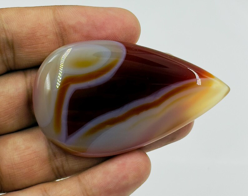 May include: A polished, teardrop-shaped cabochon gemstone featuring smooth, concentric bands of deep maroon, translucent amber, and creamy white. The stone has a high-gloss finish that reflects light, highlighting its layered, organic color patterns.