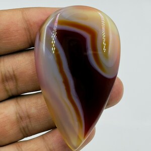 May include: A polished, teardrop-shaped agate gemstone featuring smooth, concentric bands of deep maroon, cream, and translucent amber. The stone has a glossy, reflective surface with distinct color transitions and a smooth, rounded edge profile.