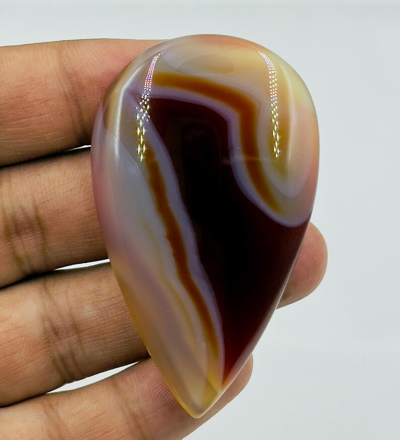 May include: A polished, teardrop-shaped agate gemstone featuring smooth, concentric bands of deep maroon, cream, and translucent amber. The stone has a glossy, reflective surface with distinct color transitions and a smooth, rounded edge profile.