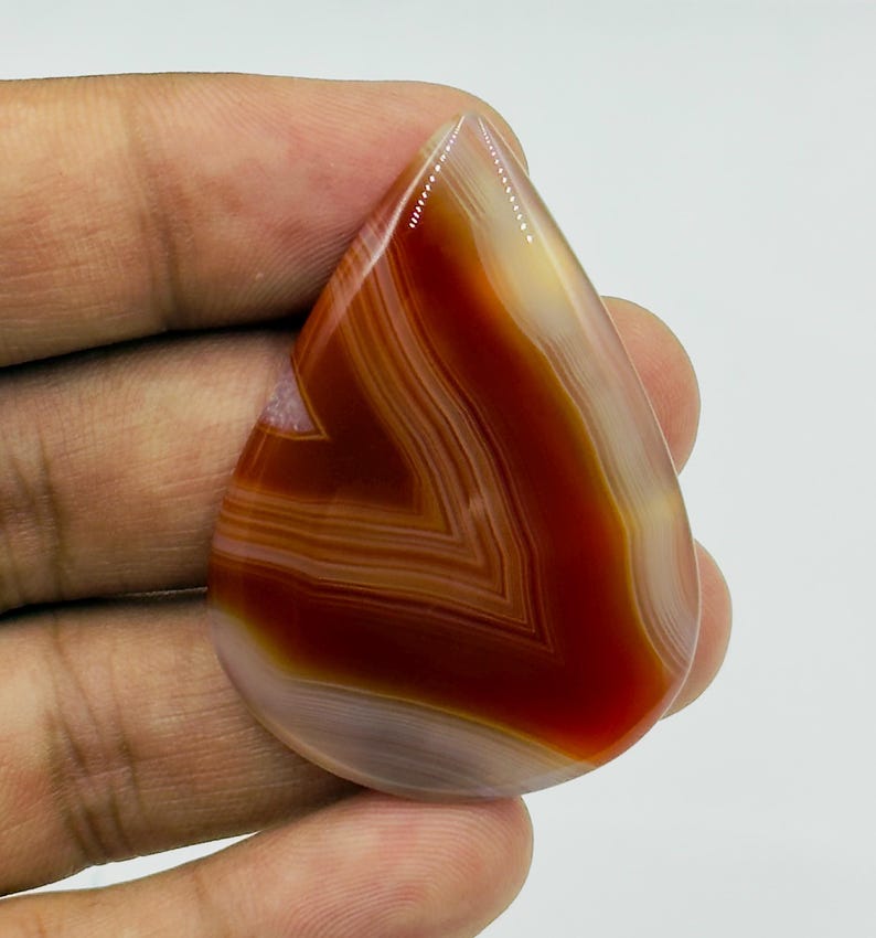 Pode incluir: Uma pedra preciosa de &aacute;gata polida em forma de l&aacute;grima com faixas conc&ecirc;ntricas de laranja queimado profundo, castanho avermelhado e branco creme transl&uacute;cido. A pedra tem uma superf&iacute;cie lisa e brilhante com um padr&atilde;o de chevron vis&iacute;vel.