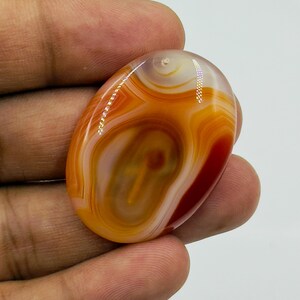 May include: An oval-shaped polished agate gemstone featuring concentric bands of orange, red, and cream colors. The stone has a smooth, glossy surface with natural swirling patterns and translucent areas that catch the light, creating a vibrant appearance.