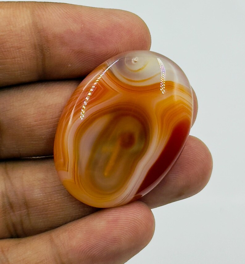 May include: An oval-shaped polished agate gemstone featuring concentric bands of orange, red, and cream colors. The stone has a smooth, glossy surface with natural swirling patterns and translucent areas that catch the light, creating a vibrant appearance.