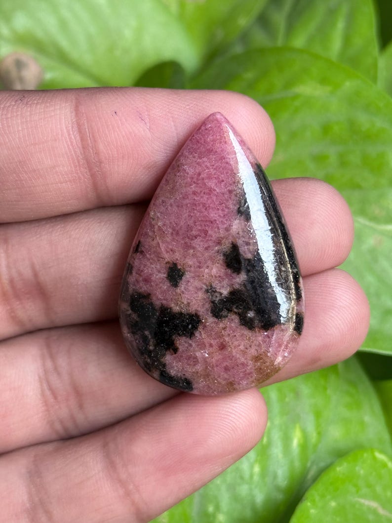 K&ouml;nnte beinhalten: Ein polierter, tropfenf&ouml;rmiger Edelstein mit rosa Grundfarbe und schwarzen und braunen Einschl&uuml;ssen. Der Stein wird in einer Hand gehalten, mit gr&uuml;nem Laub im Hintergrund. Der Edelstein hat eine gl&auml;nzende Oberfl&auml;che.