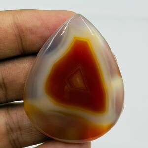 May include: A polished, teardrop-shaped agate gemstone featuring a vibrant, deep red-orange center surrounded by concentric bands of translucent white and pale yellow, showcasing a smooth, glossy surface with natural mineral banding patterns.