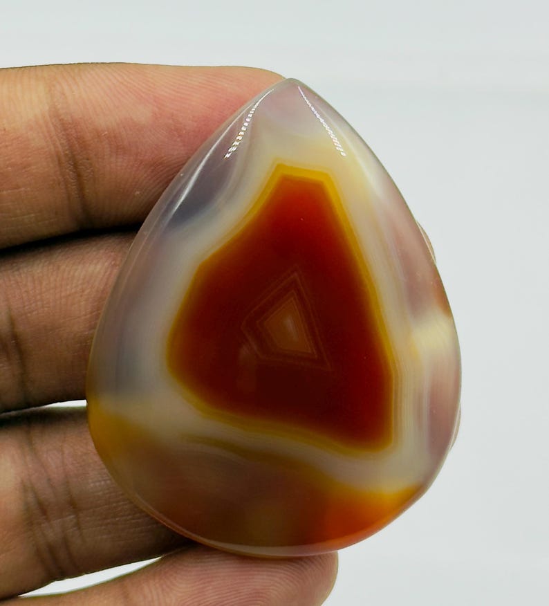 May include: A polished, teardrop-shaped agate gemstone featuring a vibrant, deep red-orange center surrounded by concentric bands of translucent white and pale yellow, showcasing a smooth, glossy surface with natural mineral banding patterns.