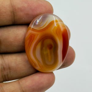 May include: An oval-shaped polished gemstone featuring concentric bands of translucent orange, deep red, and creamy white colors. The stone has a smooth, glossy surface with a distinct layered pattern radiating from the center toward the outer edges.