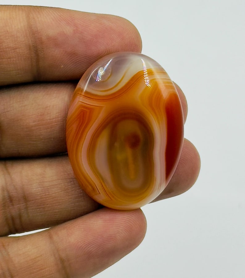 May include: An oval-shaped polished gemstone featuring concentric bands of translucent orange, deep red, and creamy white colors. The stone has a smooth, glossy surface with a distinct layered pattern radiating from the center toward the outer edges.