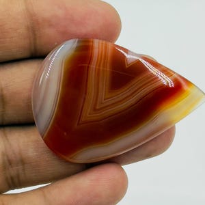 Pode incluir: Uma pedra preciosa de &aacute;gata cornalina polida em forma de l&aacute;grima, apresentando bandas conc&ecirc;ntricas distintas de laranja queimado profundo, castanho avermelhado e branco transl&uacute;cido. A pedra tem uma superf&iacute;cie lisa e brilhante que reflete a luz.