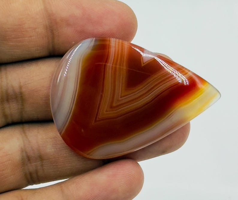Pode incluir: Uma pedra preciosa de &aacute;gata cornalina polida em forma de l&aacute;grima, apresentando bandas conc&ecirc;ntricas distintas de laranja queimado profundo, castanho avermelhado e branco transl&uacute;cido. A pedra tem uma superf&iacute;cie lisa e brilhante que reflete a luz.