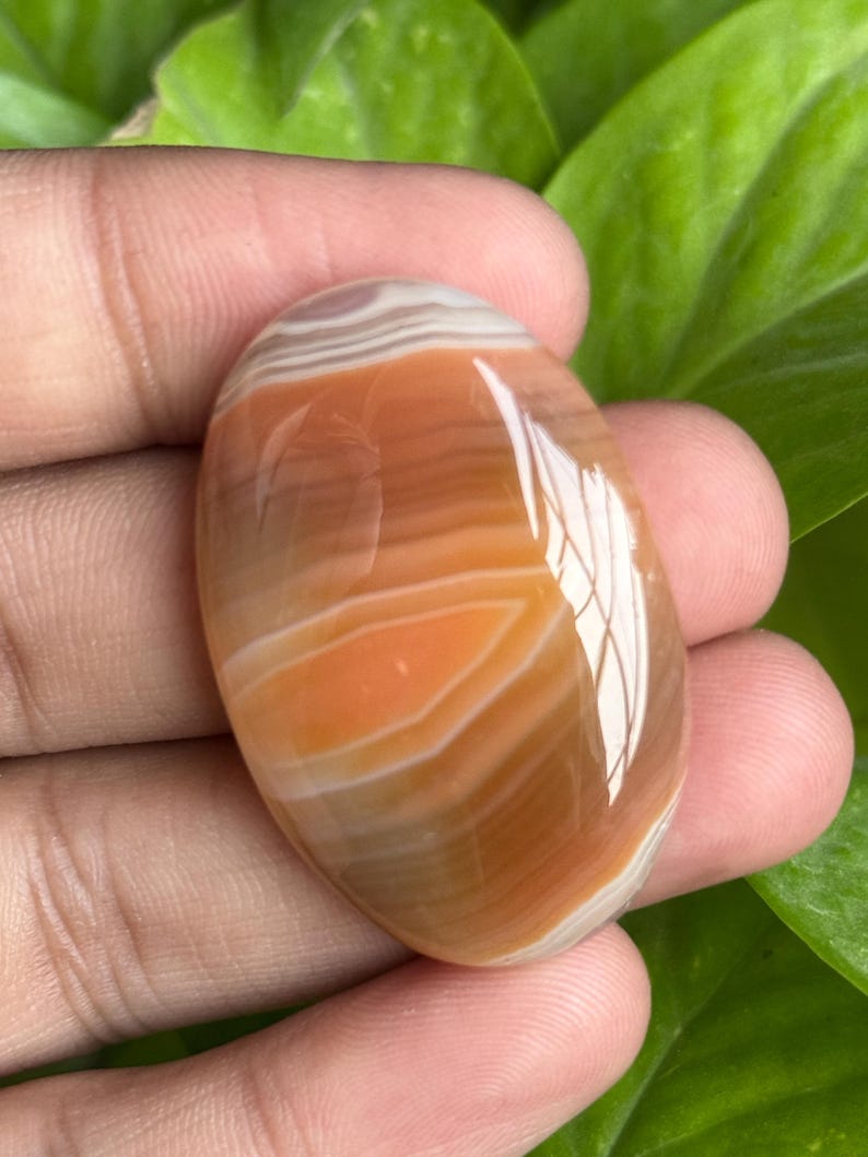 May include: Oval-shaped polished gemstone with layers of orange, brown, and white. The stone has a smooth, glossy surface and a natural, earthy appearance. The gemstone is held in a hand, with green foliage in the background.