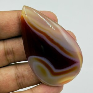 May include: A polished, teardrop-shaped agate gemstone featuring smooth, concentric bands of deep mahogany, translucent white, and soft golden yellow. The stone has a high-gloss finish that reflects light, highlighting its layered, organic color patterns.