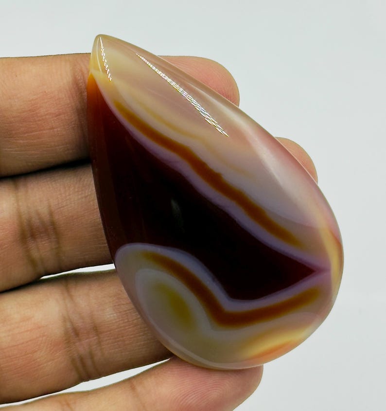 May include: A polished, teardrop-shaped agate gemstone featuring smooth, concentric bands of deep mahogany, translucent white, and soft golden yellow. The stone has a high-gloss finish that reflects light, highlighting its layered, organic color patterns.