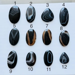 May include: Twelve polished cabochon gemstones arranged in a grid, numbered one through twelve. These stones feature black, white, and brown banded patterns in oval and teardrop shapes, displaying natural concentric layers and smooth surfaces.