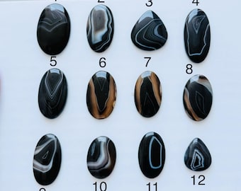 May include: Twelve polished cabochon gemstones arranged in a grid, numbered one through twelve. These stones feature black, white, and brown banded patterns in oval and teardrop shapes, displaying natural concentric layers and smooth surfaces.