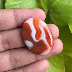 May include: An oval-shaped cabochon gemstone featuring smooth, polished layers of vibrant orange, deep reddish-brown, and opaque white banding. The stone has a glossy, reflective surface that highlights the intricate, flowing patterns within the mineral structure.