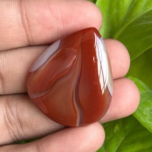May include: A polished, teardrop-shaped gemstone with a rich, reddish-brown hue, accented by swirling white and cream-colored bands. The stone is held in a hand, showcasing its smooth, reflective surface and natural patterns. The gemstone is a natural stone.