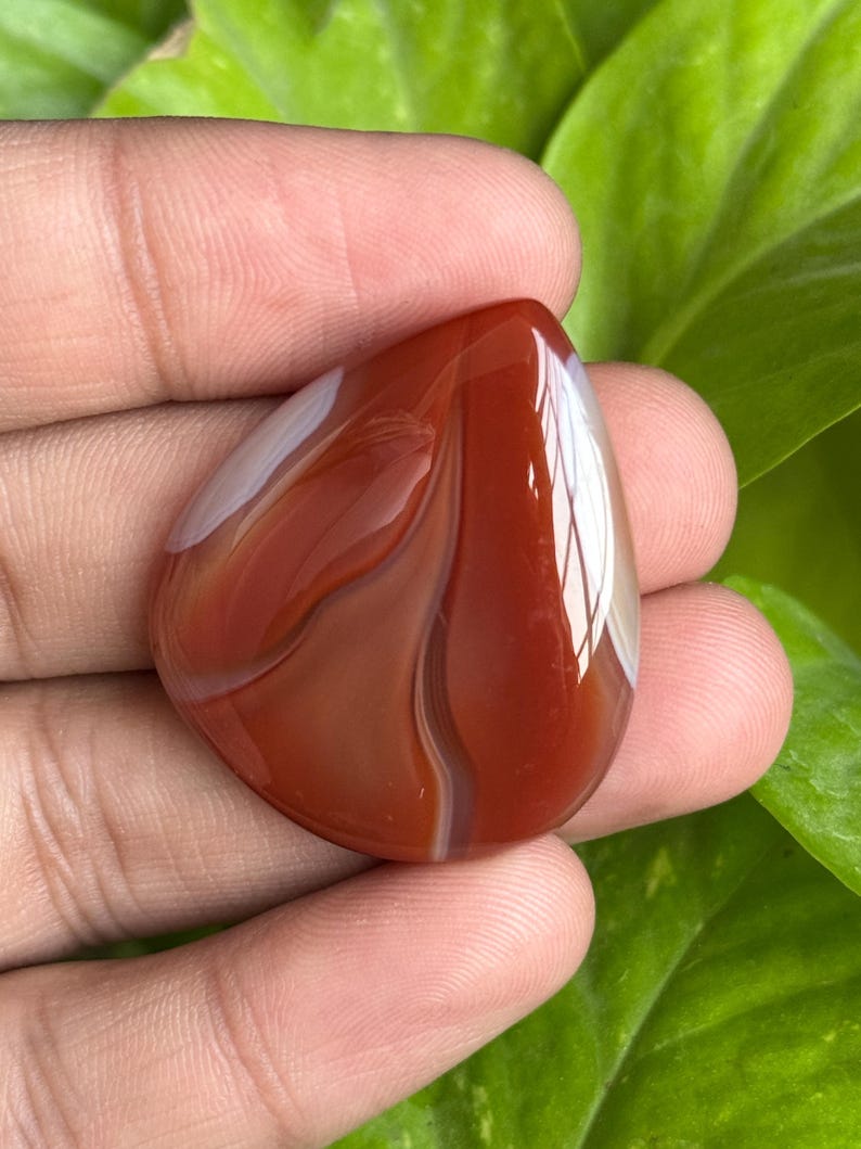 May include: A polished, teardrop-shaped gemstone with a rich, reddish-brown hue, accented by swirling white and cream-colored bands. The stone is held in a hand, showcasing its smooth, reflective surface and natural patterns. The gemstone is a natural stone.