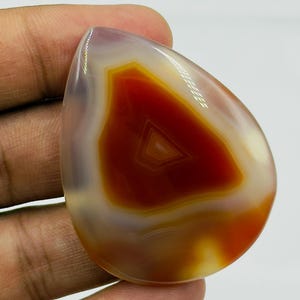 May include: A polished, teardrop-shaped agate gemstone featuring a vibrant, deep red center with concentric banding patterns. The stone transitions into translucent, milky white and soft grey edges, showcasing a smooth, glossy surface texture.