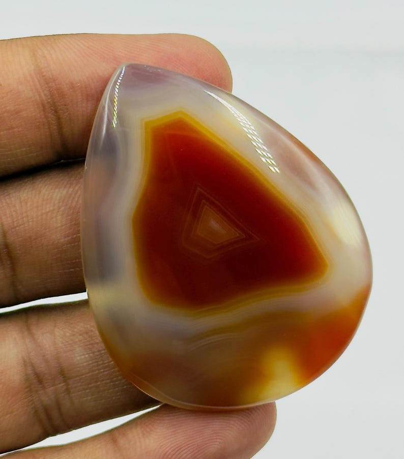 May include: A polished, teardrop-shaped agate gemstone featuring a vibrant, deep red center with concentric banding patterns. The stone transitions into translucent, milky white and soft grey edges, showcasing a smooth, glossy surface texture.