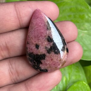 K&ouml;nnte beinhalten: Ein polierter, tropfenf&ouml;rmiger Edelstein mit einem rosa-schwarzen Muster. Der Stein hat eine gl&auml;nzende Oberfl&auml;che und wird in einer Hand vor einem Hintergrund aus gr&uuml;nen Bl&auml;ttern gehalten. Die rosa Farbe variiert in den Schattierungen, und das Schwarz erscheint in unregelm&auml;&szlig;igen Flecken.