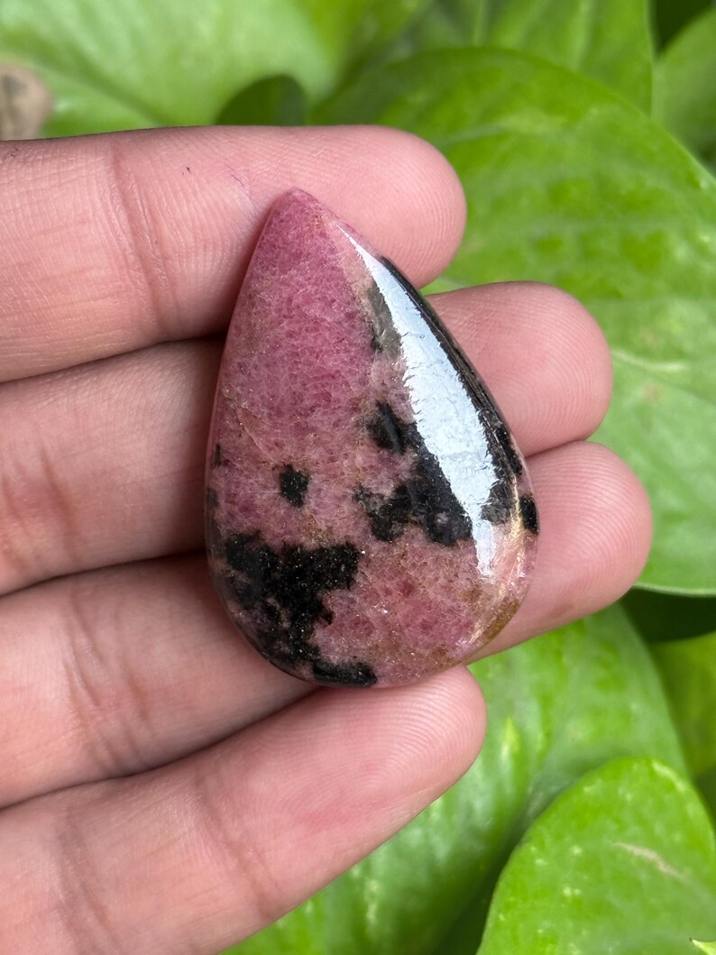 K&ouml;nnte beinhalten: Ein polierter, tropfenf&ouml;rmiger Edelstein mit einem rosa-schwarzen Muster. Der Stein hat eine gl&auml;nzende Oberfl&auml;che und wird in einer Hand vor einem Hintergrund aus gr&uuml;nen Bl&auml;ttern gehalten. Die rosa Farbe variiert in den Schattierungen, und das Schwarz erscheint in unregelm&auml;&szlig;igen Flecken.