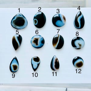 May include: A collection of twelve polished, smooth cabochon gemstones arranged in three rows on a white surface. Each stone features distinct black, white, and light blue banded patterns in various shapes, including teardrops and circles, numbered 1 to 12.