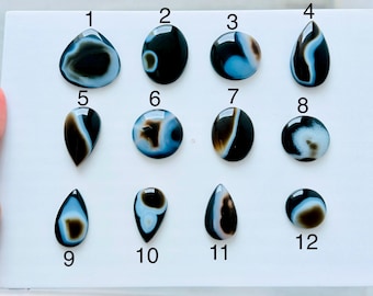 May include: A collection of twelve polished, smooth cabochon gemstones arranged in three rows on a white surface. Each stone features distinct black, white, and light blue banded patterns in various shapes, including teardrops and circles, numbered 1 to 12.