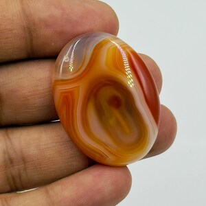May include: An oval-shaped polished gemstone featuring concentric bands of translucent orange, deep red, and creamy white colors. The stone has a smooth, glossy surface with visible light reflections on its curved, rounded top and edges.