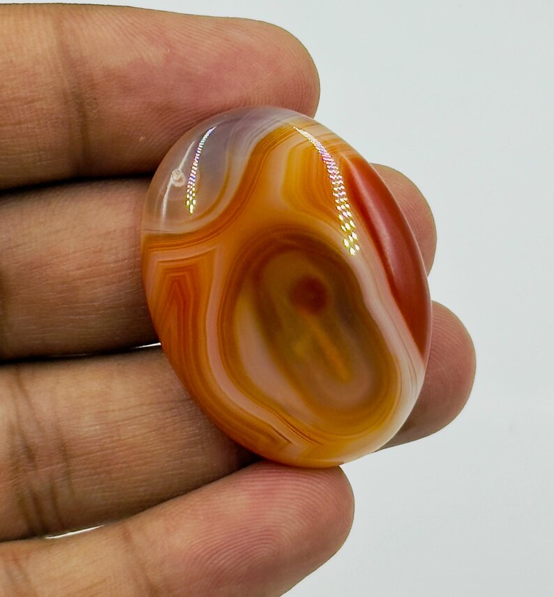 May include: An oval-shaped polished gemstone featuring concentric bands of translucent orange, deep red, and creamy white colors. The stone has a smooth, glossy surface with visible light reflections on its curved, rounded top and edges.
