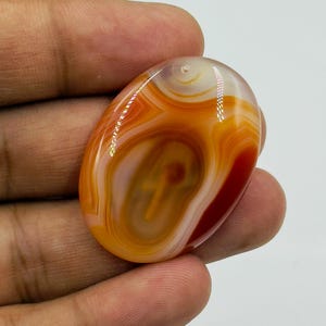 May include: An oval-shaped polished carnelian agate gemstone featuring concentric bands of translucent orange, deep red, and creamy white colors. The stone has a smooth, glossy surface with a small drilled hole at the top for suspension.