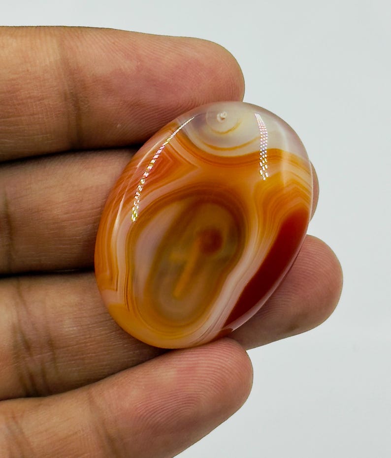 May include: An oval-shaped polished carnelian agate gemstone featuring concentric bands of translucent orange, deep red, and creamy white colors. The stone has a smooth, glossy surface with a small drilled hole at the top for suspension.