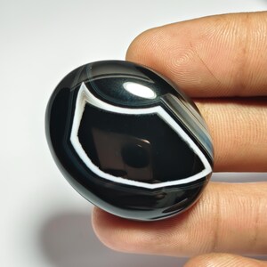 May include: A polished, oval-shaped black agate gemstone with a prominent white and gray banded pattern. The stone is held in a hand, showcasing its smooth, reflective surface and natural design. The gemstone's size is approximately 1.5 inches in length.