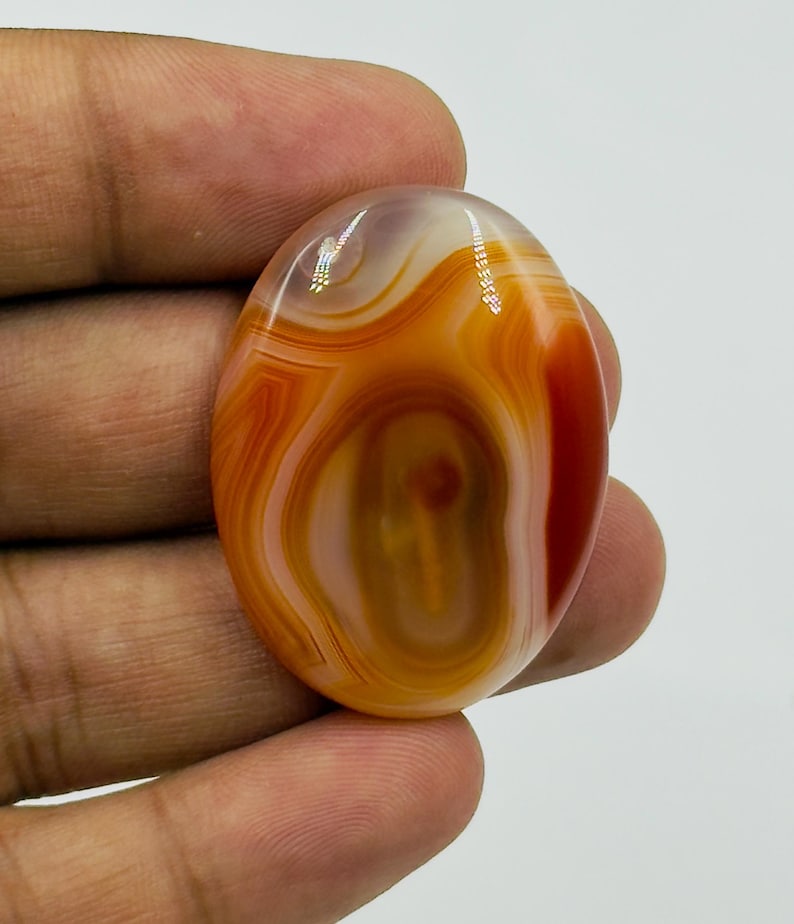 May include: An oval-shaped cabochon gemstone featuring concentric bands of translucent orange, deep red, and creamy white colors. The polished stone displays a smooth, glossy surface with natural swirling patterns and light reflections.