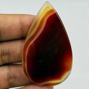 May include: A polished, teardrop-shaped agate gemstone featuring a deep, dark red center that transitions into vibrant orange and creamy white concentric bands. The stone has a smooth, glossy surface with a translucent quality at the edges.