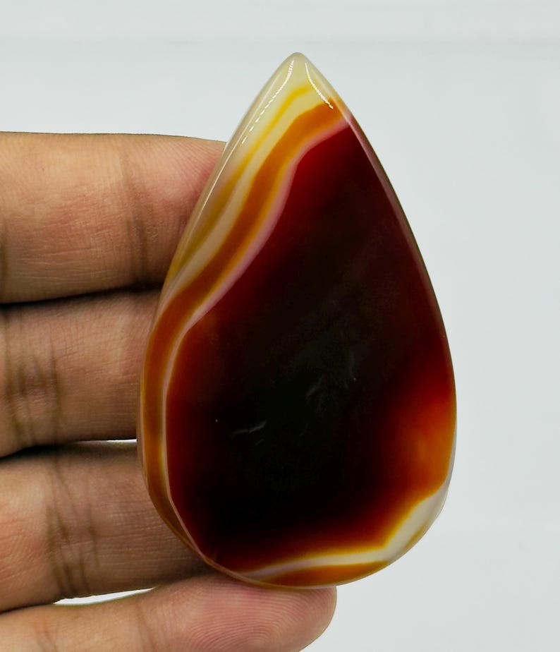 May include: A polished, teardrop-shaped agate gemstone featuring a deep, dark red center that transitions into vibrant orange and creamy white concentric bands. The stone has a smooth, glossy surface with a translucent quality at the edges.
