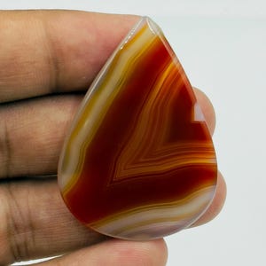 Pode incluir: Uma pedra preciosa de &aacute;gata polida em forma de l&aacute;grima com faixas conc&ecirc;ntricas de vermelho profundo, laranja queimado e branco creme. A pedra tem uma superf&iacute;cie lisa e brilhante com uma qualidade transl&uacute;cida, exibindo padr&otilde;es naturais.