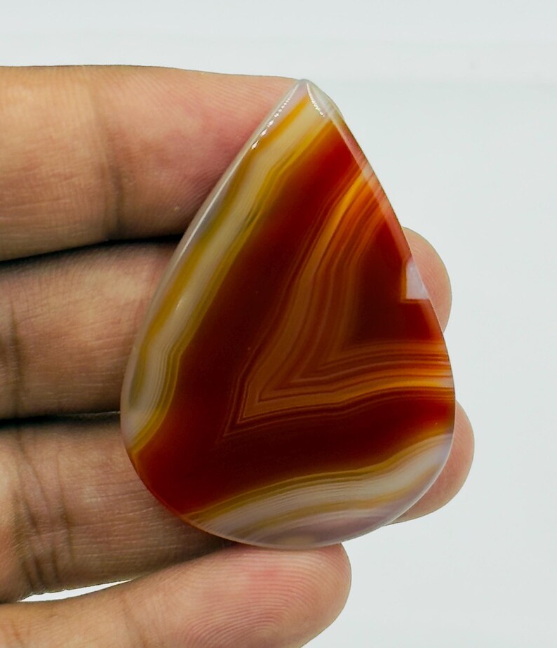 Pode incluir: Uma pedra preciosa de &aacute;gata polida em forma de l&aacute;grima com faixas conc&ecirc;ntricas de vermelho profundo, laranja queimado e branco creme. A pedra tem uma superf&iacute;cie lisa e brilhante com uma qualidade transl&uacute;cida, exibindo padr&otilde;es naturais.