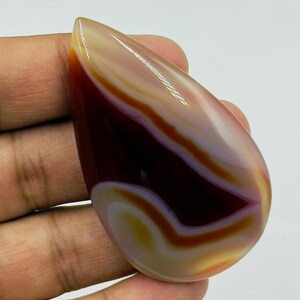 May include: A polished, teardrop-shaped cabochon gemstone featuring smooth, concentric bands of deep mahogany, translucent amber, and creamy white. The stone has a glossy, reflective surface with distinct color transitions and a curved, organic form.