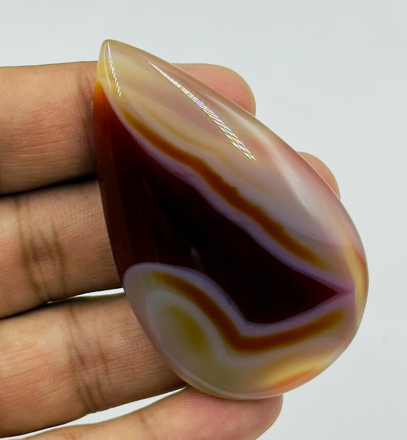 May include: A polished, teardrop-shaped cabochon gemstone featuring smooth, concentric bands of deep mahogany, translucent amber, and creamy white. The stone has a glossy, reflective surface with distinct color transitions and a curved, organic form.