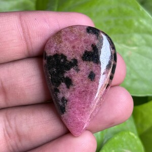 K&ouml;nnte beinhalten: Ein polierter, tropfenf&ouml;rmiger Edelstein mit einem rosa und schwarzen Muster. Der Stein hat eine gl&auml;nzende Oberfl&auml;che und wird in einer Hand vor einem Hintergrund aus gr&uuml;nem Laub gehalten. Die rosa Farbe dominiert, mit schwarzen Einschl&uuml;ssen.
