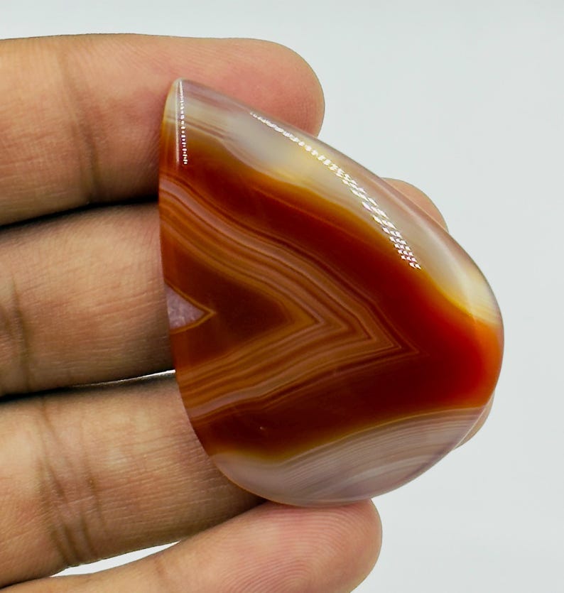 Pode incluir: Uma pedra preciosa de &aacute;gata polida em forma de l&aacute;grima com bandas conc&ecirc;ntricas distintas de laranja queimado, vermelho ferrugem e branco transl&uacute;cido. A superf&iacute;cie lisa e brilhante reflete a luz, destacando os padr&otilde;es minerais.