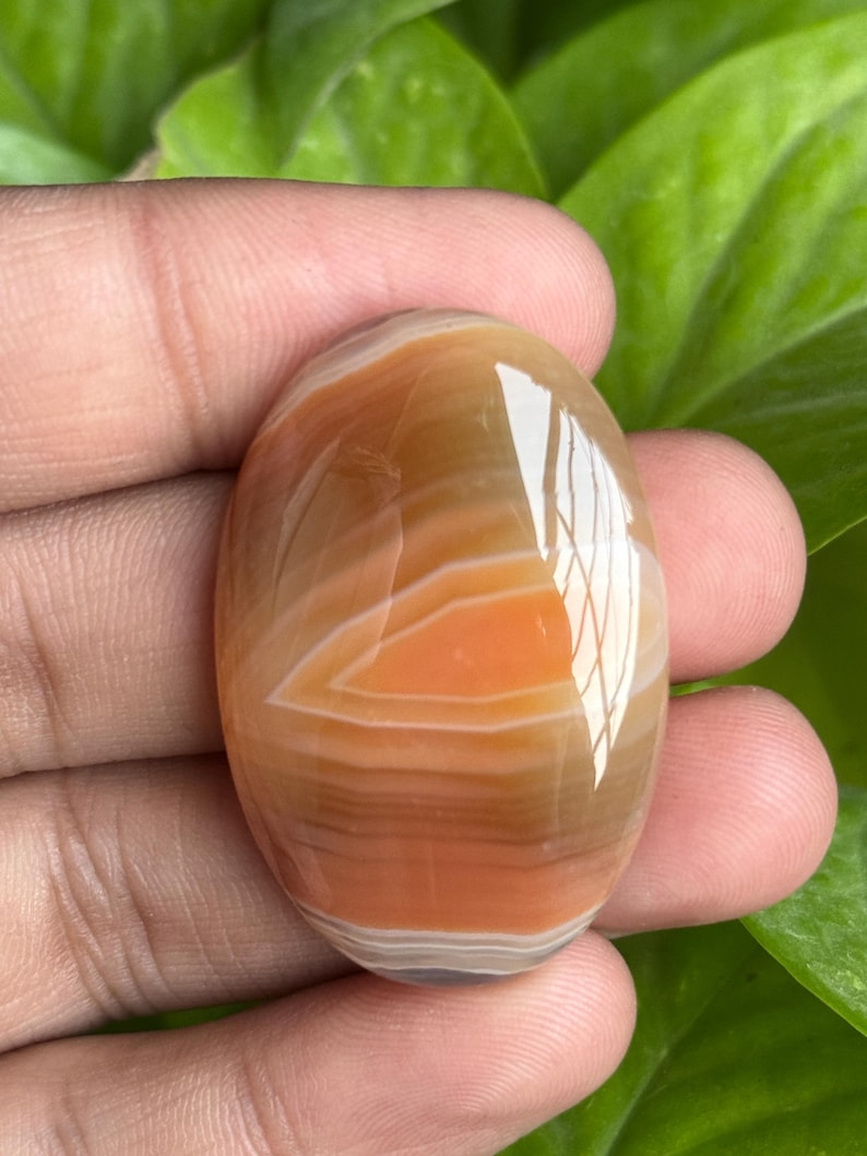 May include: Oval-shaped polished gemstone with bands of orange, brown, and white. The stone has a glossy finish and is held in a hand, with green foliage in the background. The gemstone is approximately 2 inches long.