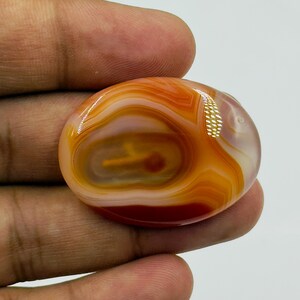 May include: An oval-shaped, polished carnelian gemstone featuring concentric bands of translucent orange, deep red, and creamy white colors. The stone has a smooth, glossy surface with a distinct layered pattern and a bright light reflection.