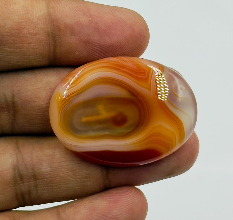 May include: An oval-shaped, polished carnelian gemstone featuring concentric bands of translucent orange, deep red, and creamy white colors. The stone has a smooth, glossy surface with a distinct layered pattern and a bright light reflection.