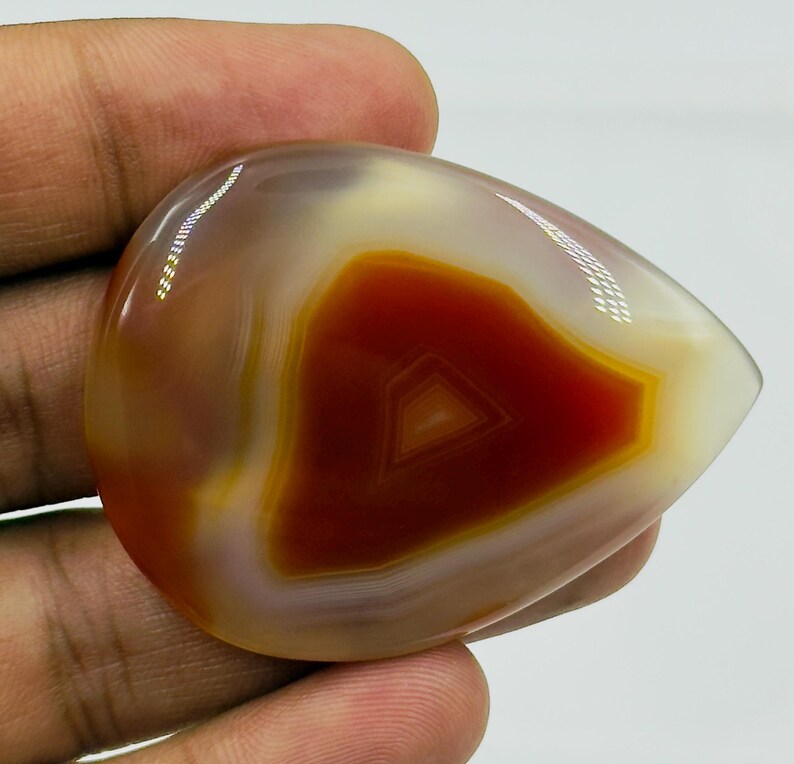 May include: A polished, teardrop-shaped agate gemstone featuring concentric bands of deep burnt orange, vibrant amber, and translucent cream. The stone has a smooth, glossy surface that reflects light, highlighting its natural layered mineral structure.