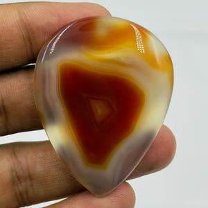 May include: A polished, teardrop-shaped cabochon gemstone featuring a vibrant central core of deep reddish-brown surrounded by concentric bands of translucent amber, golden yellow, and creamy white, with a smooth, glossy, reflective surface.